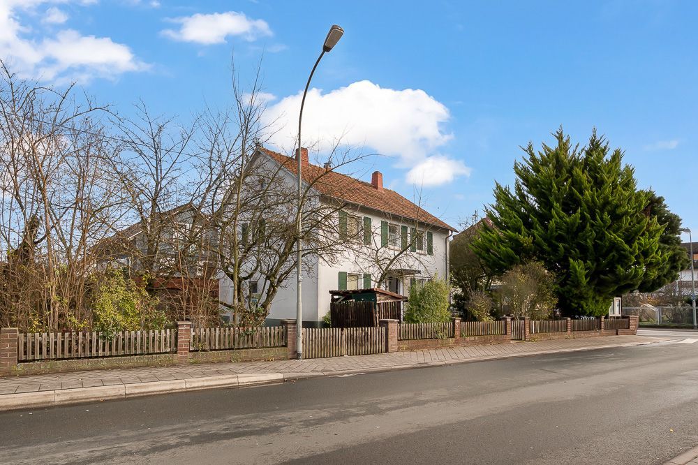 Zweistöckiges weißes Haus mit Garten und Zaun an einer Straße bei blauem Himmel.