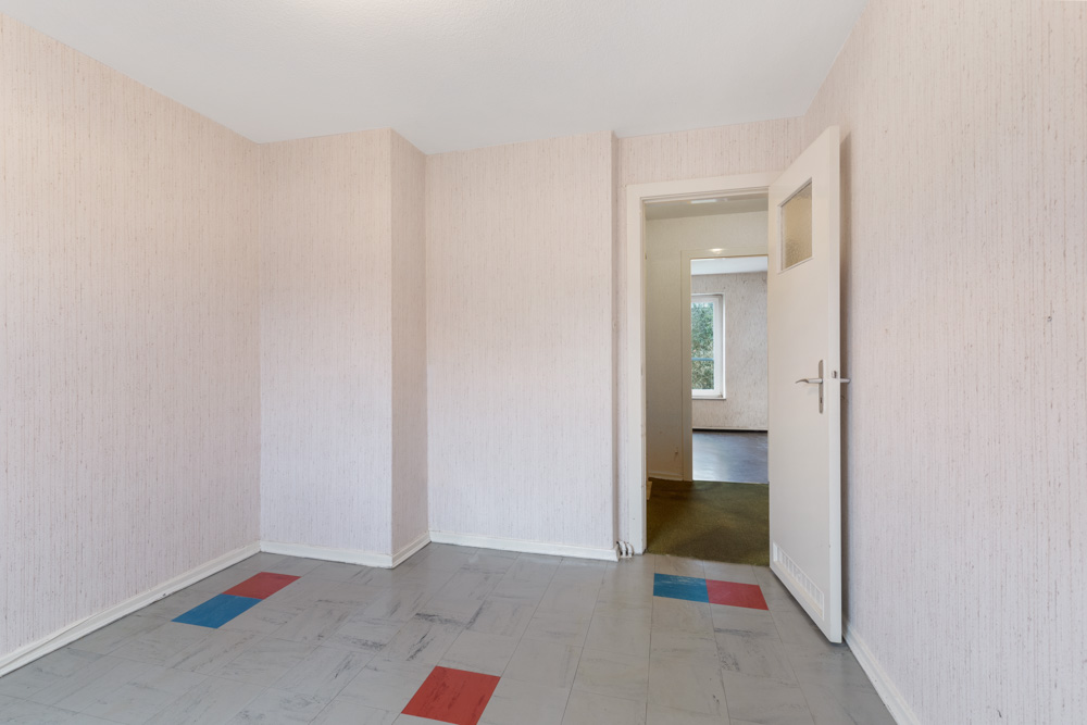 Leerer Raum mit gemustertem Boden, offenem Türrahmen und sichtbarem Fenster im Nachbarraum.