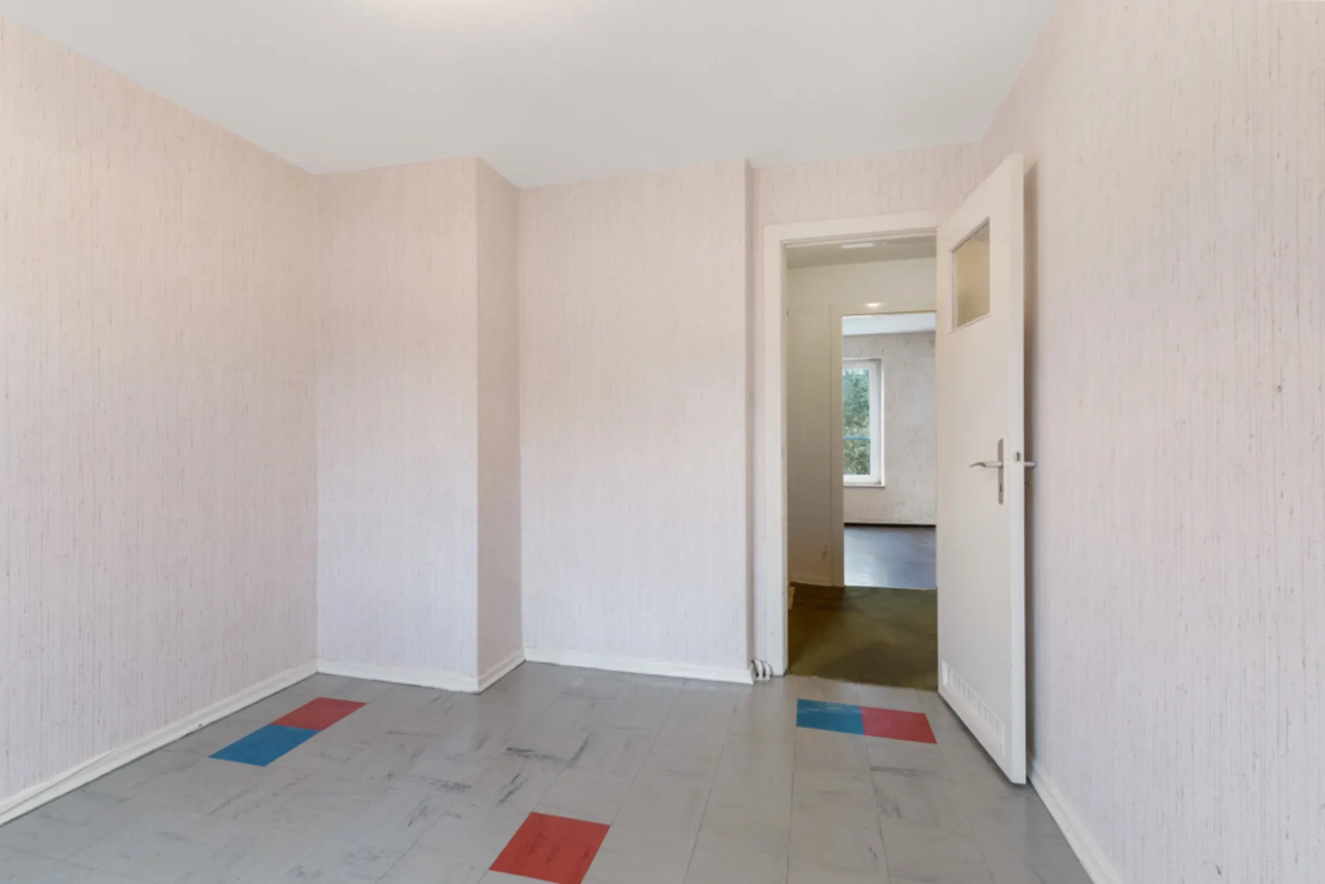 Leerer Raum mit gemustertem Boden, offenem Türrahmen und sichtbarem Fenster im Nachbarraum.