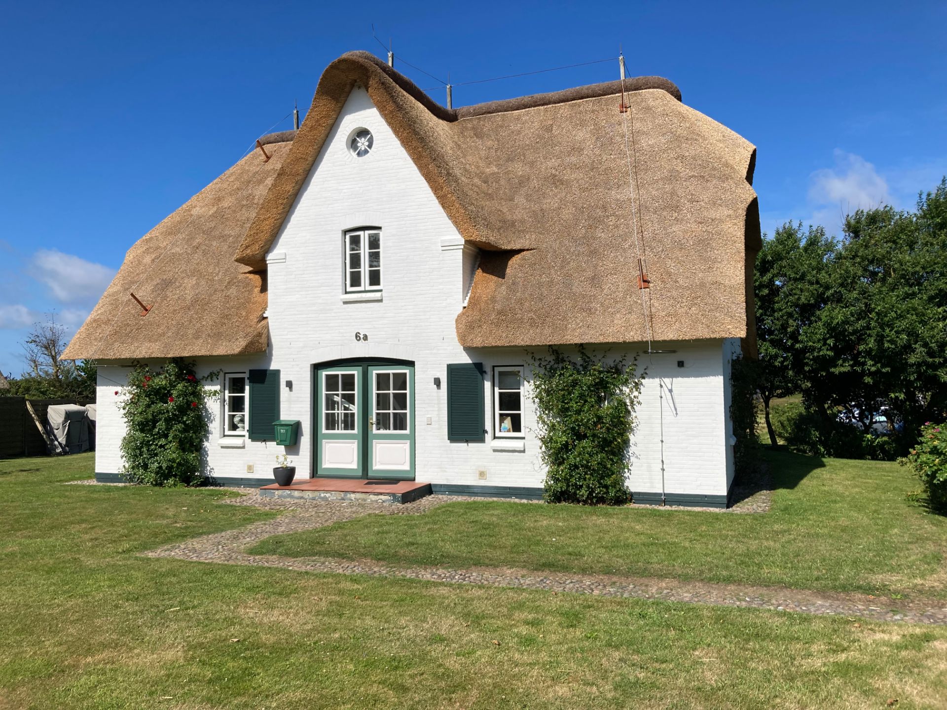 Weißes Haus mit Reetdach, grünen Fensterläden und Tür, umgeben von Rasen und Büschen.