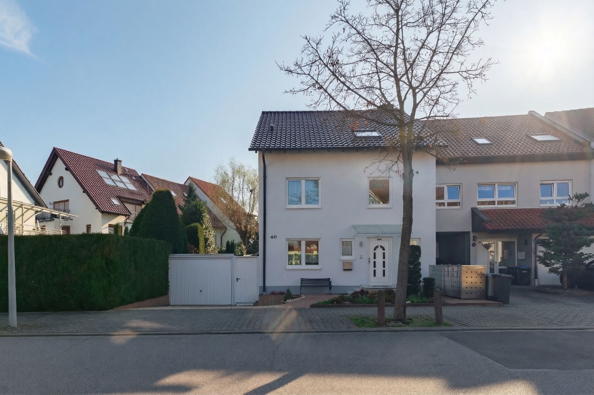 Weißes Reihenhaus mit braunem Dach, kleinem Garten und Baum davor bei sonnigem Wetter.