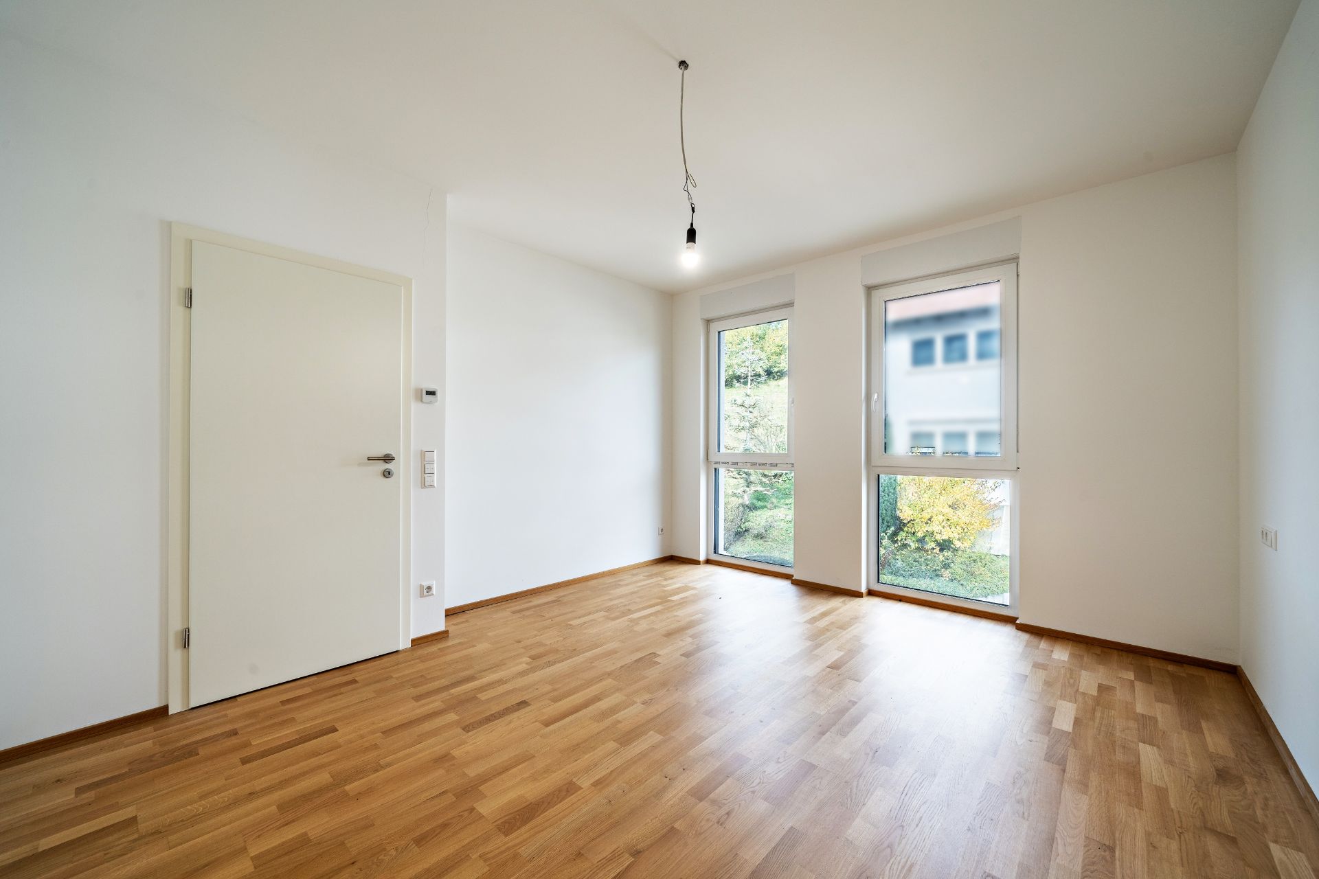 Leerer Raum mit Holzfußboden, weißer Tür und zwei großen Fenstern.