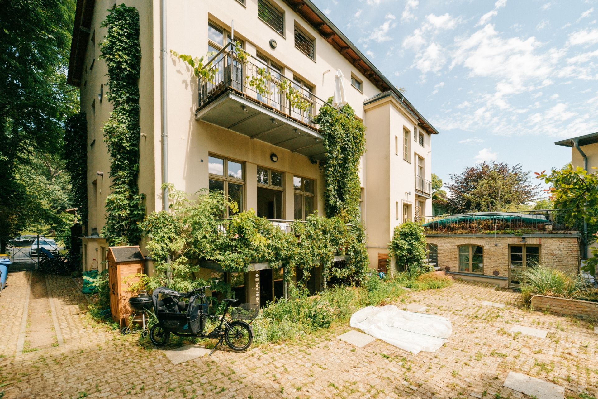 Dreistöckiges Wohnhaus mit viel grünem Bewuchs, Innenhof mit Fahrrad und Kopfsteinpflaster.