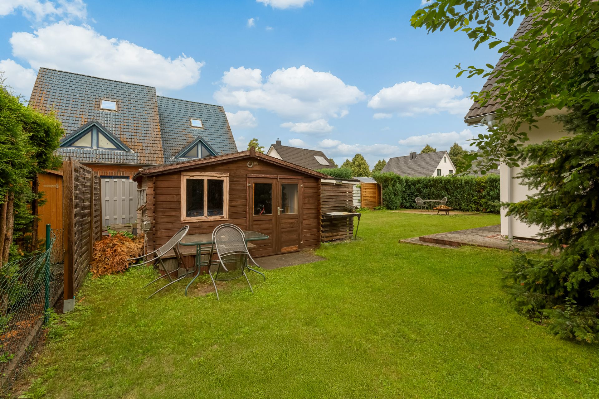 Garten mit Rasen, Holzhütte und Gartentisch mit Stühlen, umgeben von Häusern und Hecken.