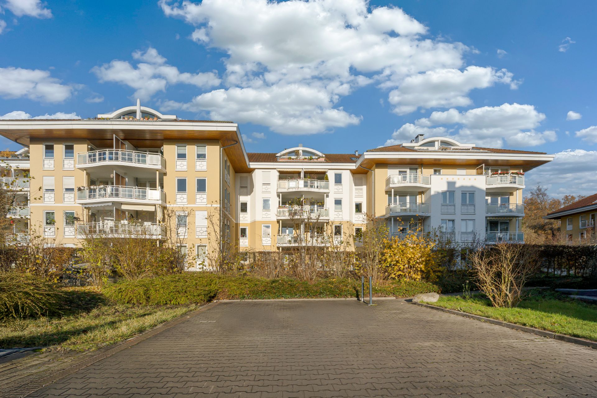 Mehrfamilienhaus mit Balkonen, umgeben von Büschen und Bäumen unter blauem Himmel.