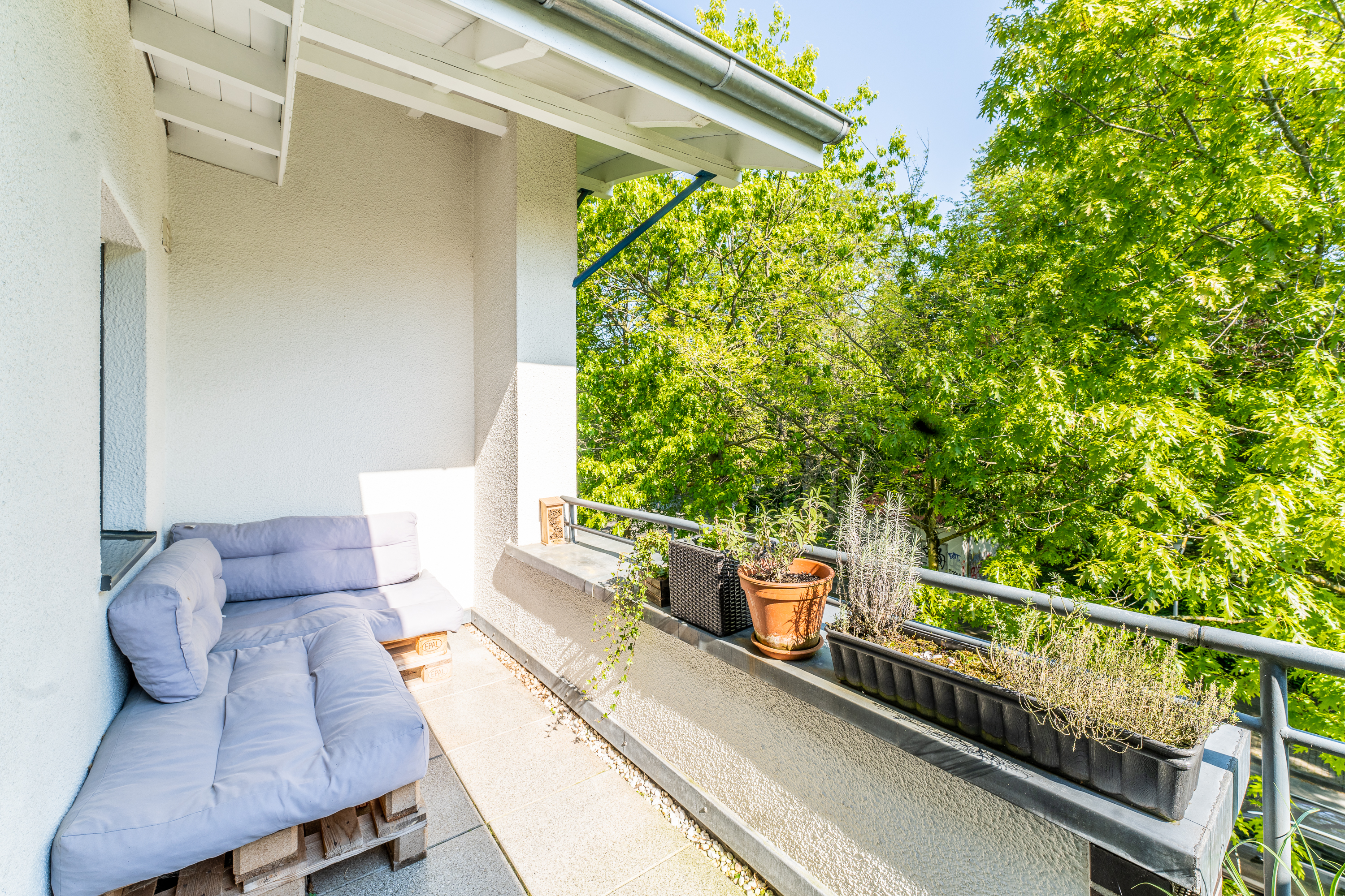 Balkon mit Palettensofa und mehreren Pflanzgefäßen am Geländer, umgeben von grünen Bäumen.
