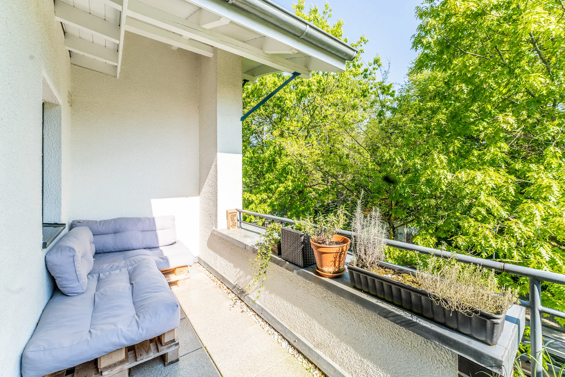 Balkon mit Palettensofa und mehreren Pflanzgefäßen am Geländer, umgeben von grünen Bäumen.