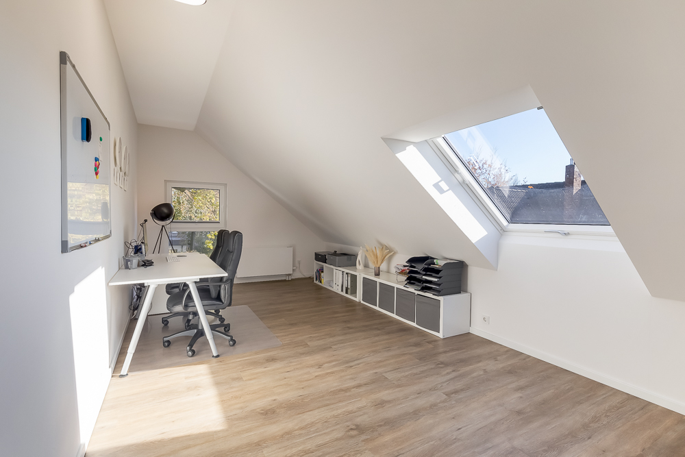 Helles Arbeitszimmer mit Dachfenster, Schreibtisch, Stuhl und Regalen auf Laminatboden.