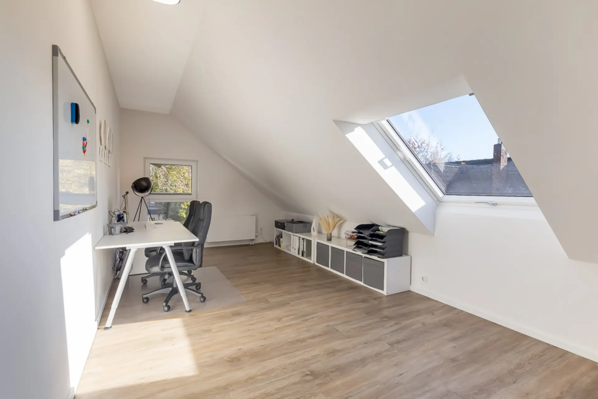 Helles Arbeitszimmer mit Dachfenster, Schreibtisch, Stuhl und Regalen auf Laminatboden.