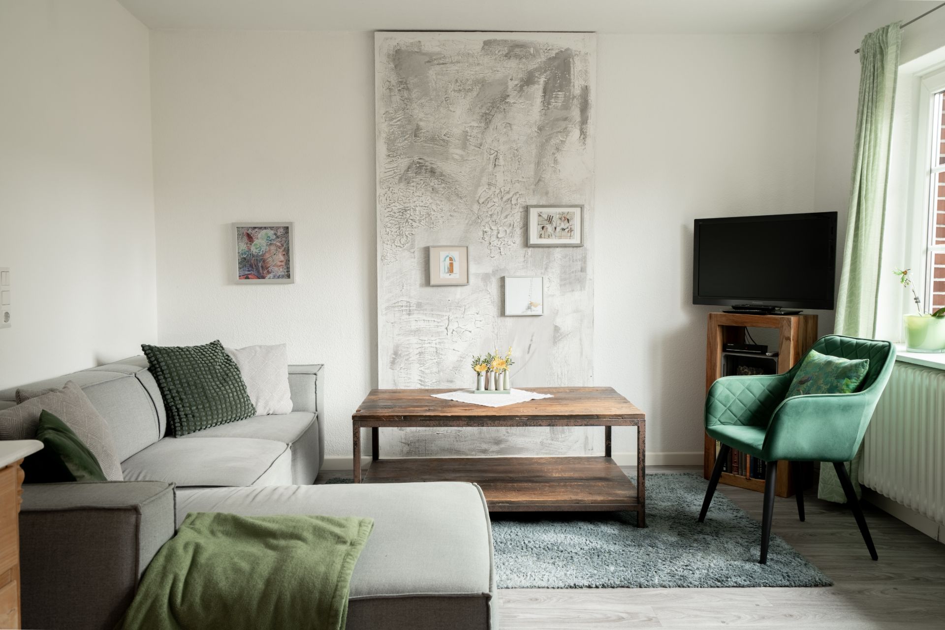 Gemütliches Wohnzimmer mit Ecksofa, grünem Sessel, Holztisch und Fernseher am Fenster.