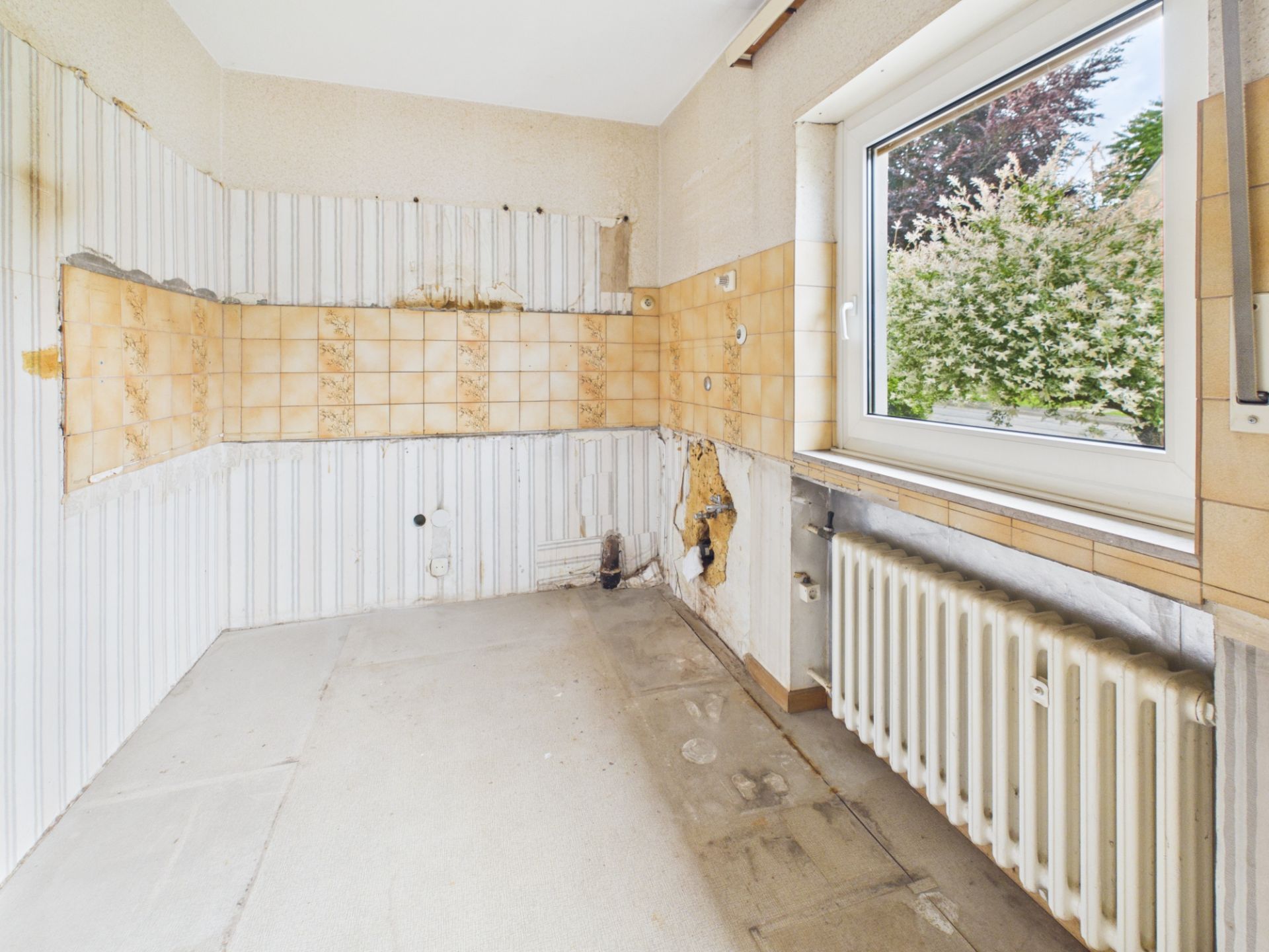 Leerer Raum mit alten Fliesen, großem Fenster und Heizkörper, sichtbare Gebrauchsspuren.