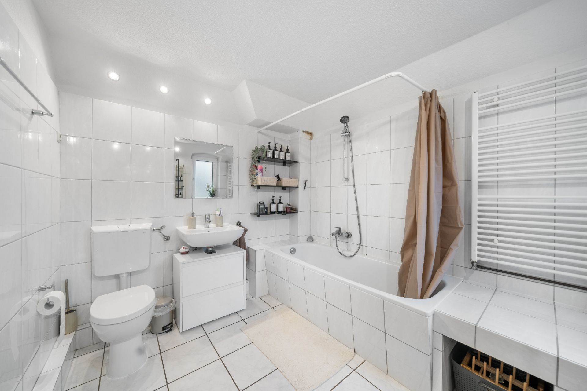Helles Bad mit Badewanne, WC, Waschbecken, Spiegel, Regal und Handtuchheizung.