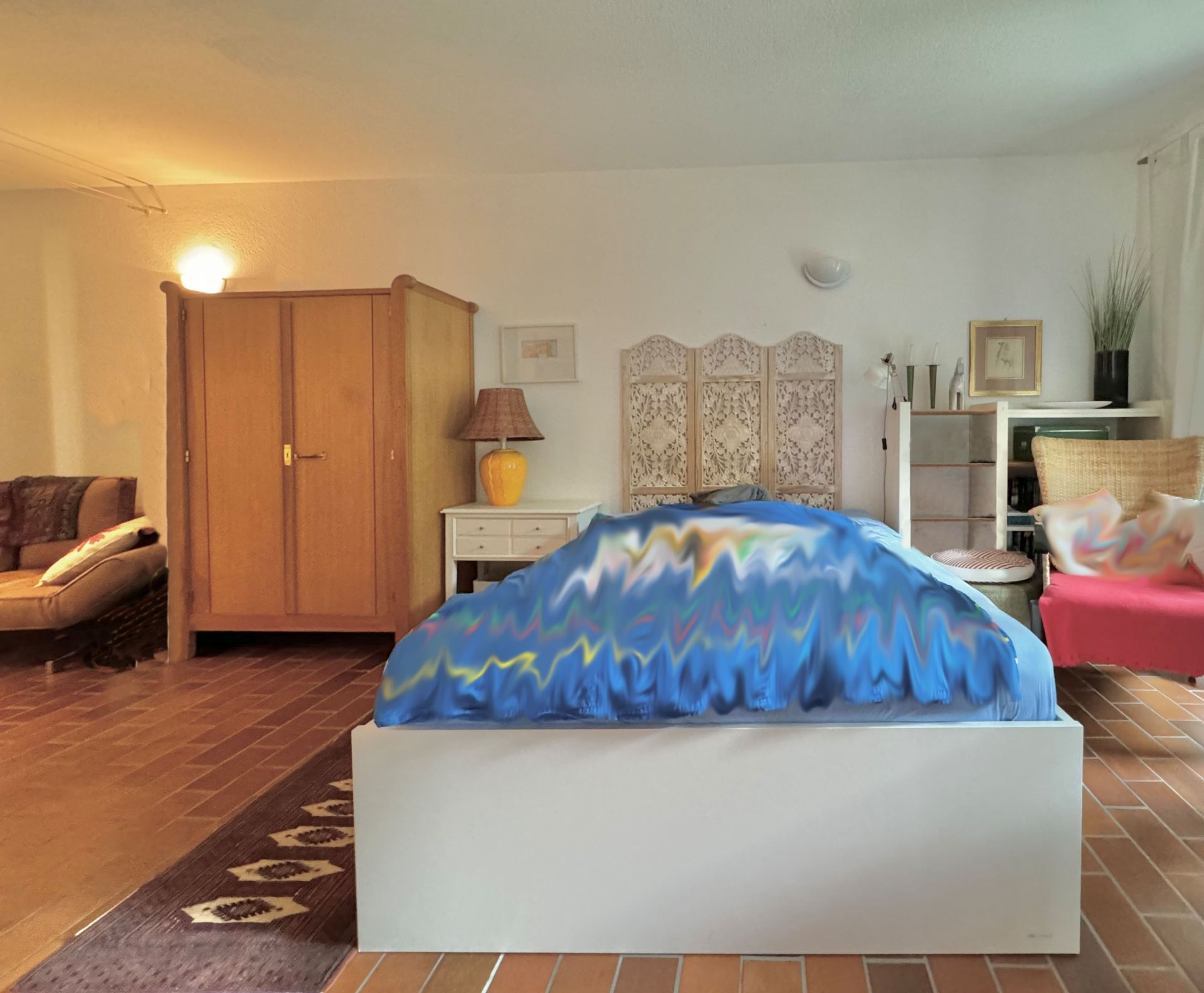 Schlafzimmer mit Doppelbett, Holzschrank, Nachttischlampe und Sitzbereich auf Fliesenboden.