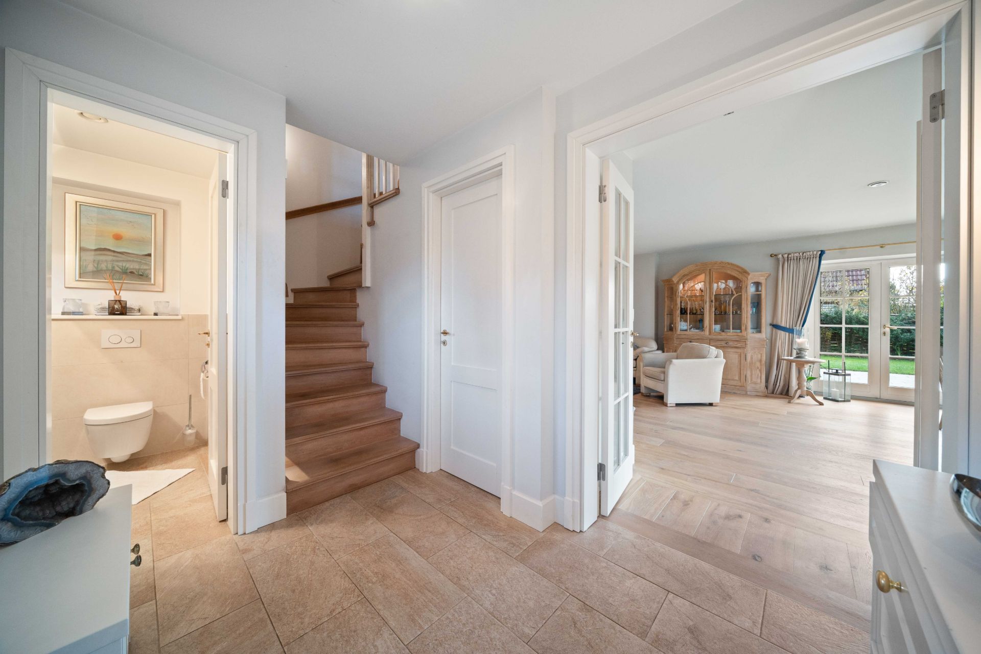 Flur mit Zugang zu einem Badezimmer, einer Holztreppe und einem hellen Wohnzimmer mit großen Fenstern.