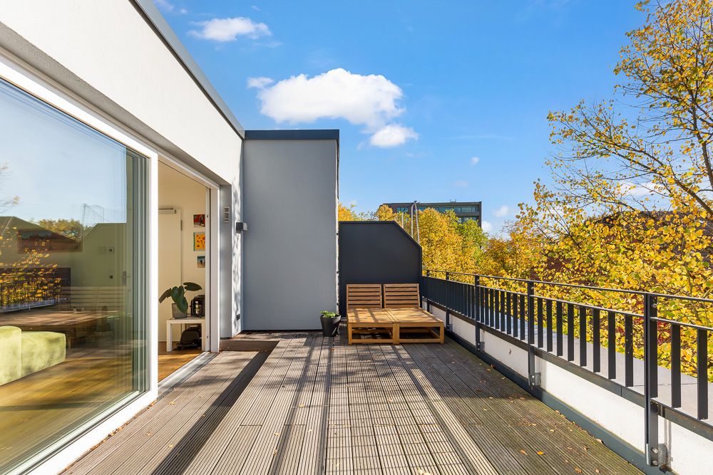 Große Terrasse mit Holzboden, Liege und Geländer, neben Wohnraum mit Schiebetür, Bäume im Hintergrund.