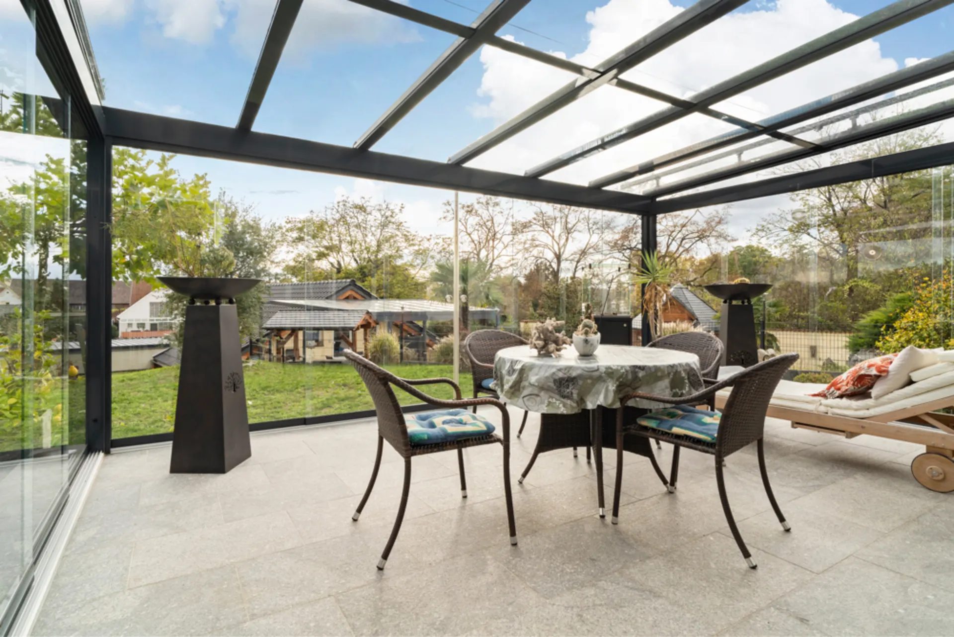 Wintergarten mit Glasdach, runder Tisch und vier Stühlen, Liege und Blick in den Garten.