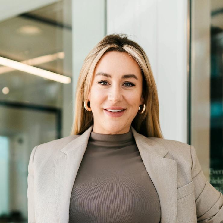 Eine Frau mit langen blonden Haaren trägt einen beigen Blazer und steht in einem modernen Innenraum.