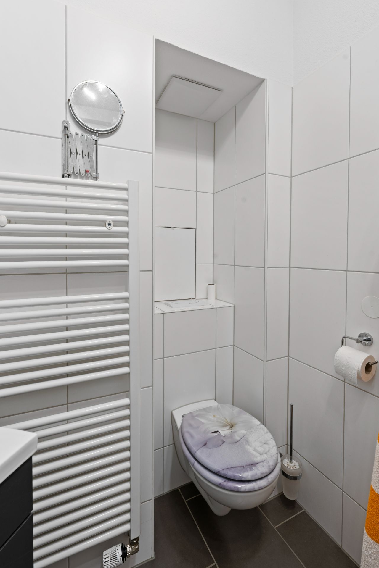 Modernes, weiß gefliestes Badezimmer mit Wandtoilette, Handtuchheizkörper und Klopapierhalter.