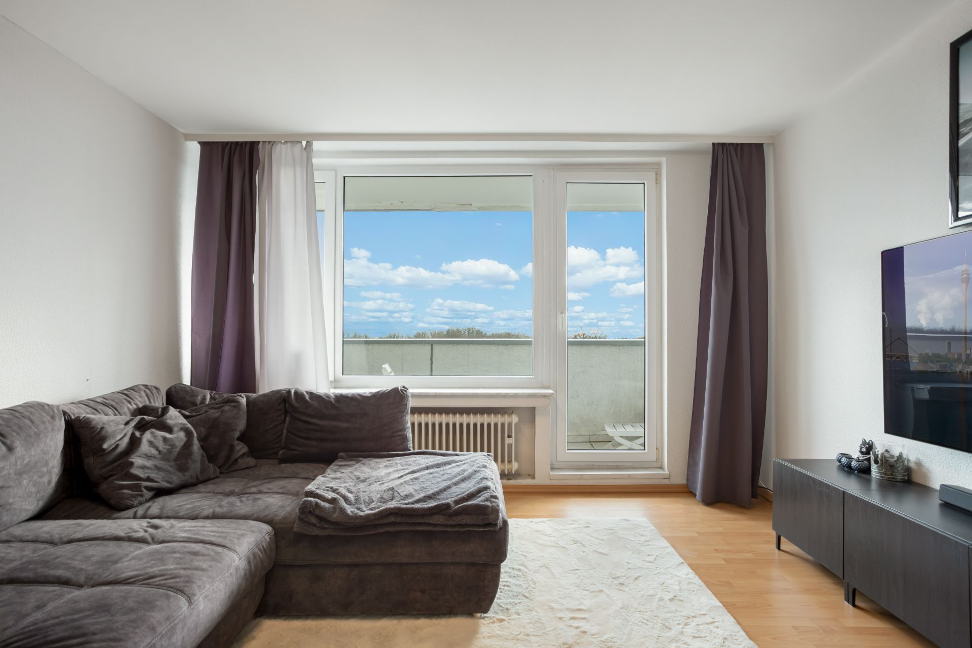 Wohnzimmer mit grauem Sofa, Fernseher und Fenster mit Blick auf Balkon und Himmel.