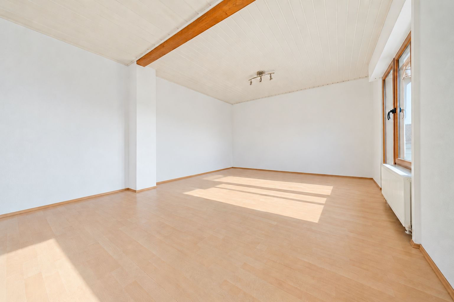 Leerer, heller Raum mit Holzfußboden, weißem Wänden und großem Fenster.