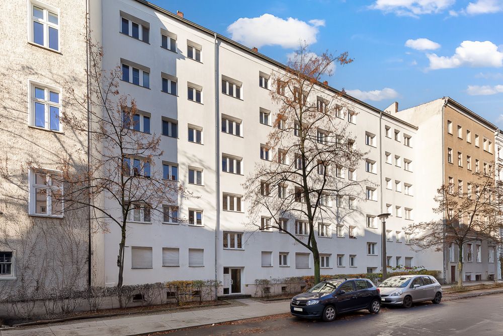 Mehrstöckiges Wohnhaus mit weißen Fassaden, Bäumen davor und geparkten Autos an der Straße.