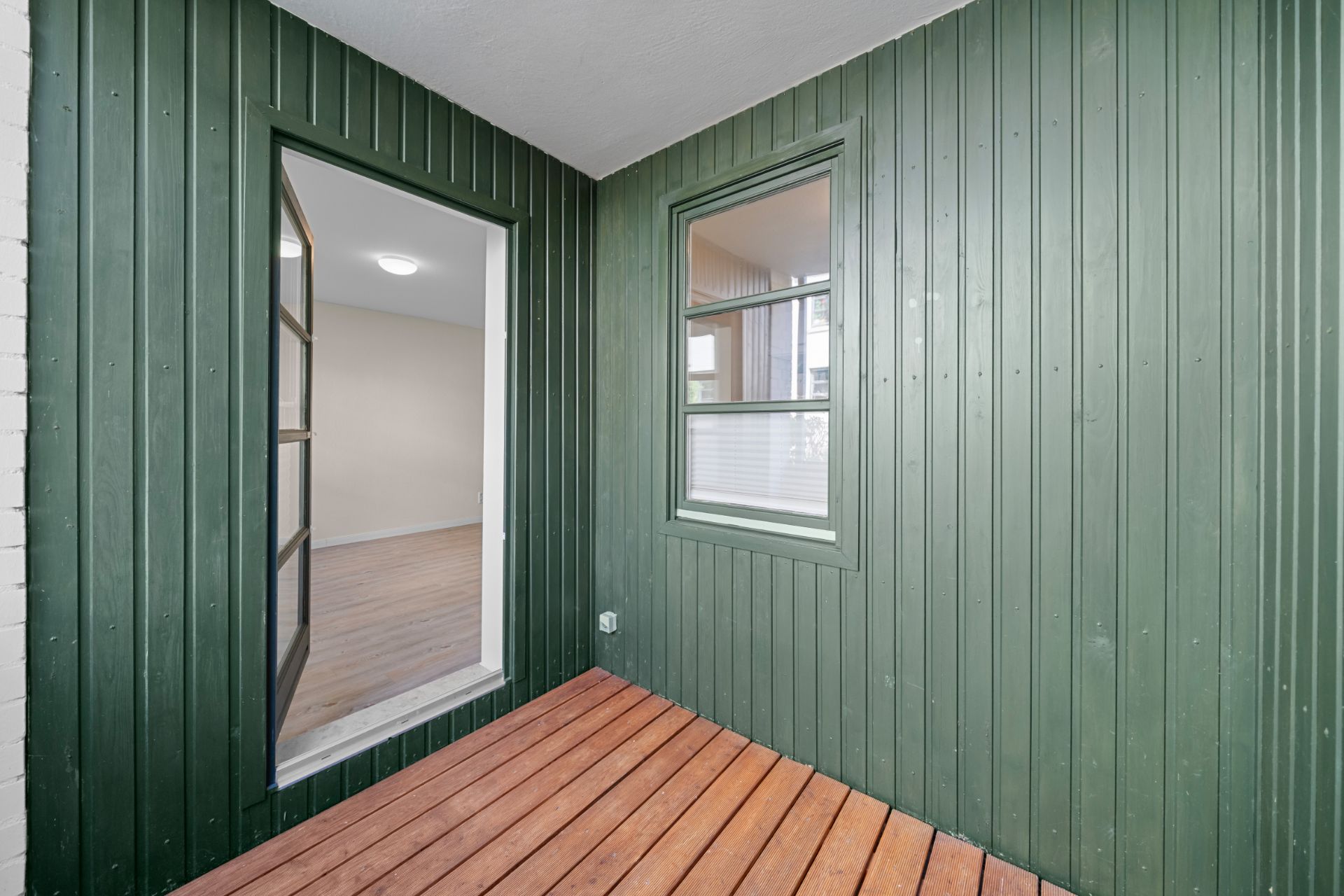 Kleiner Balkon mit grüner Holzverkleidung, Holzfußboden und Fenster zum Innenraum.