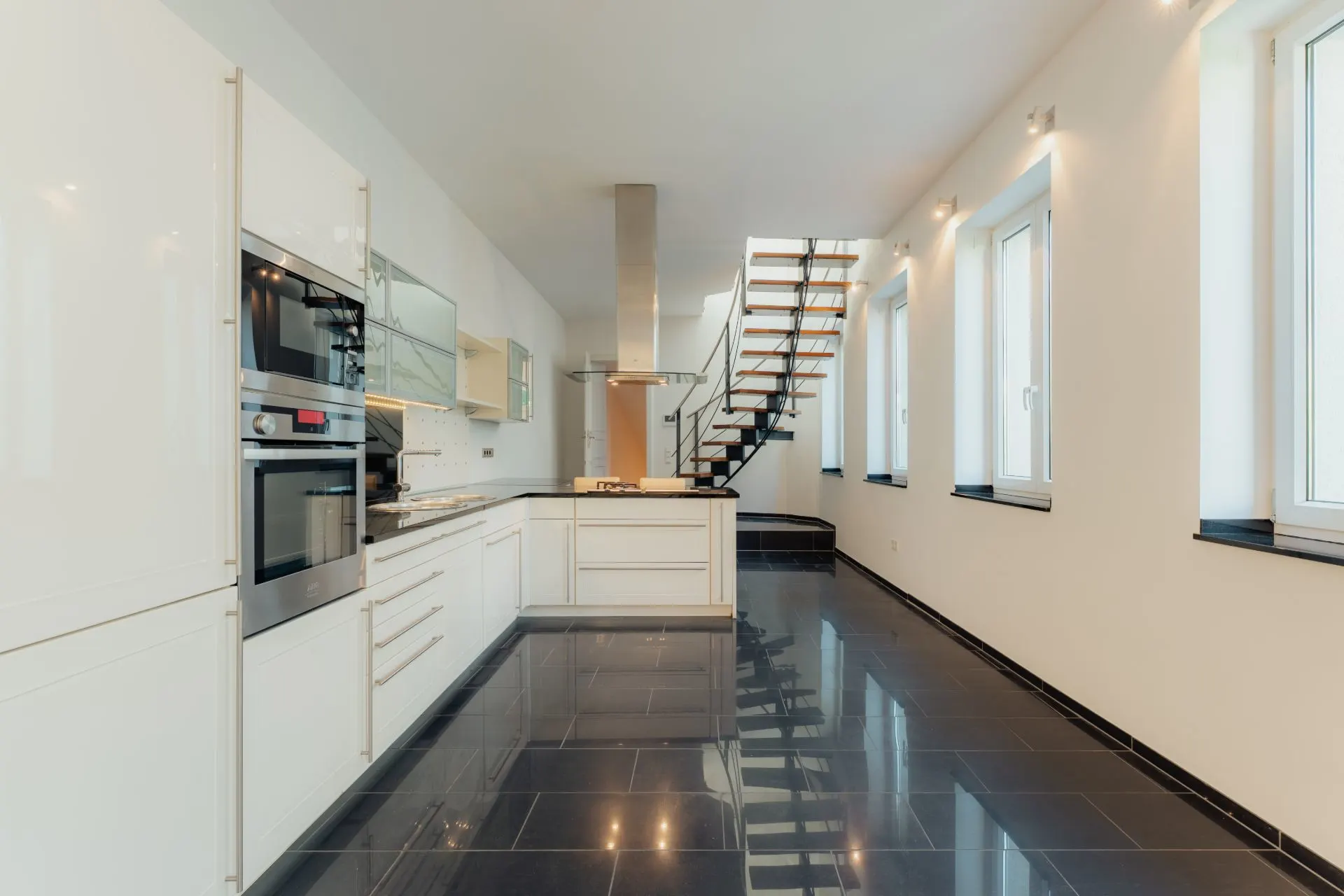 Moderne weiße Küche mit schwarzem Fliesenboden und offener Treppe, mehrere Fenster an der rechten Wand.