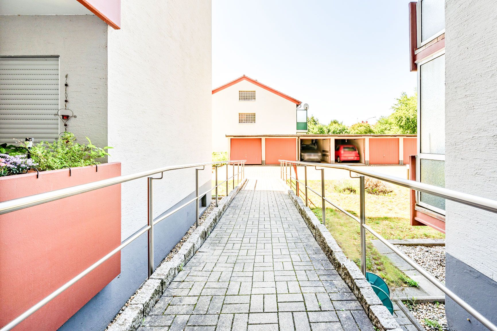 Gepflasterter Weg mit Geländer zwischen Wohngebäuden, im Hintergrund Garagen mit Autos.