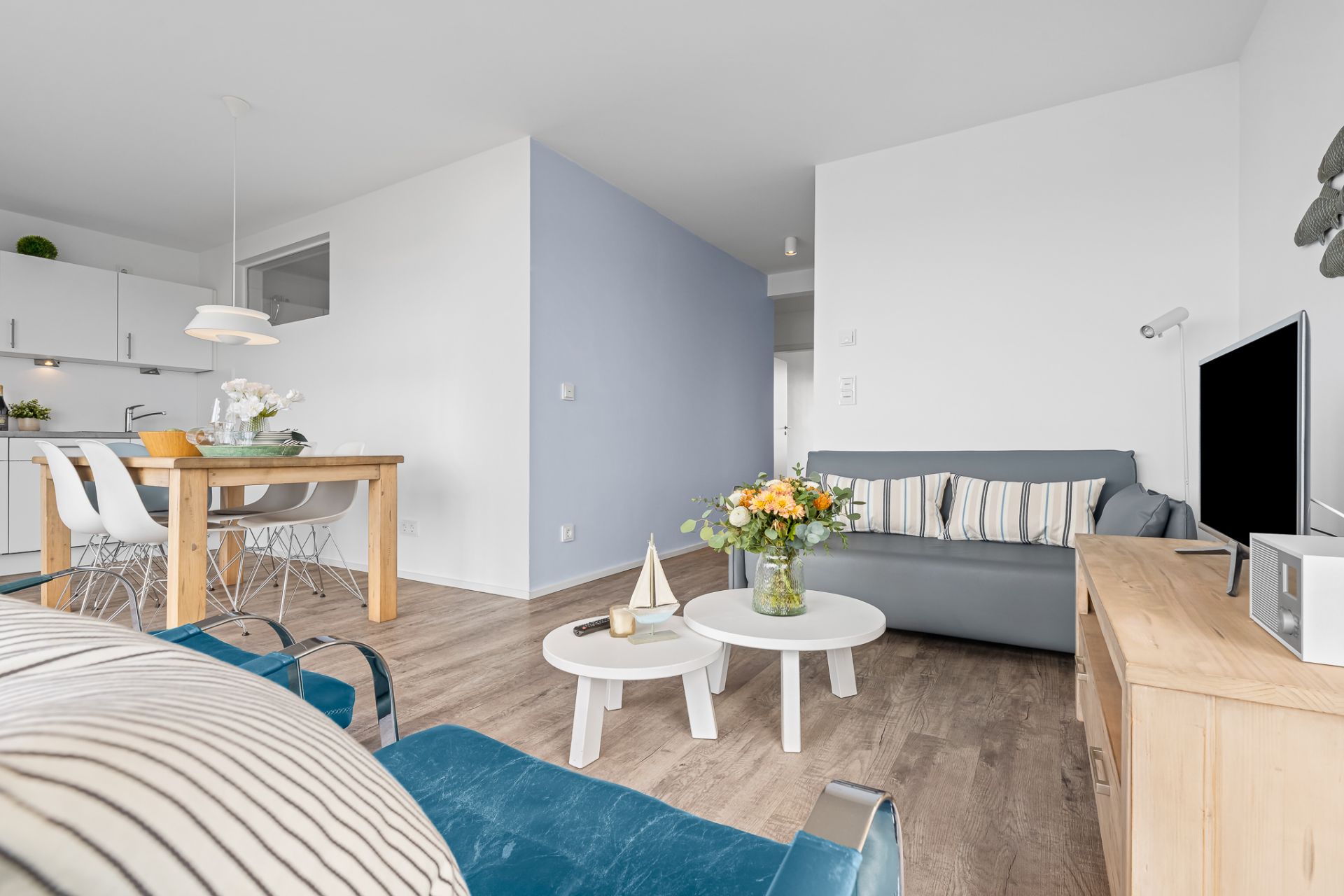Helles Wohnzimmer mit Sofa, Couchtischen, TV und offener Küche mit Esstisch.