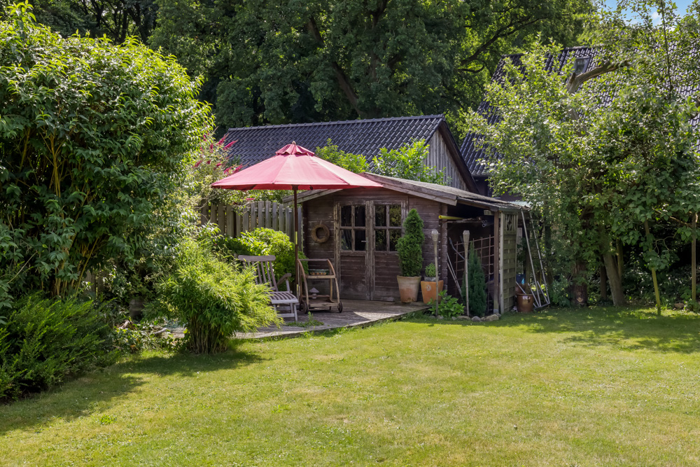 Garten mit Holzhaus, rotem Sonnenschirm und Sitzbereich, umgeben von viel Grün.