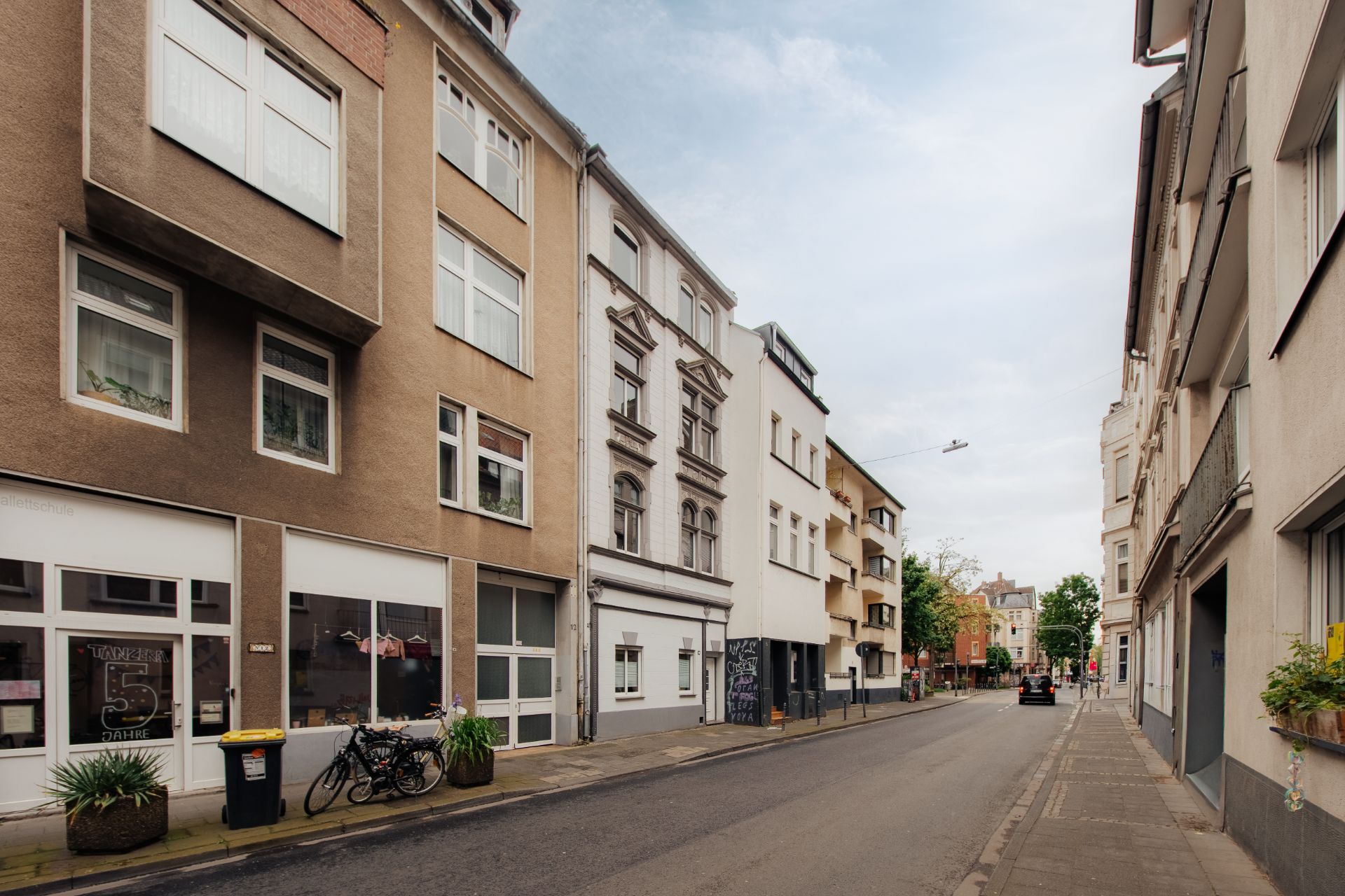 Wohnstraße mit mehrstöckigen Häusern, Gehweg, Fahrrädern und Mülleimer.