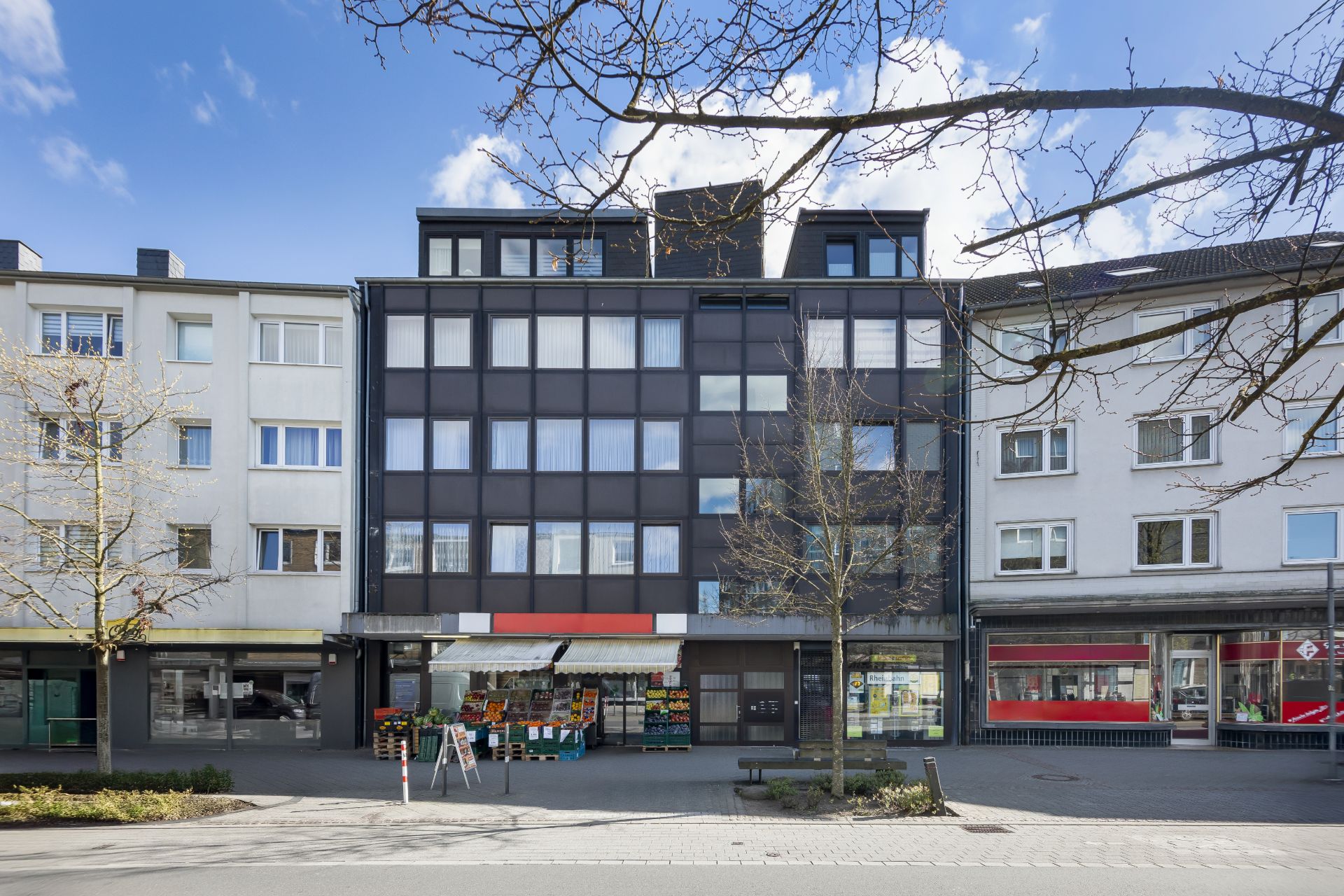 Mehrstöckiges Wohnhaus mit Laden im Erdgeschoss und großen Fenstern, davor Bäume und Gehweg.