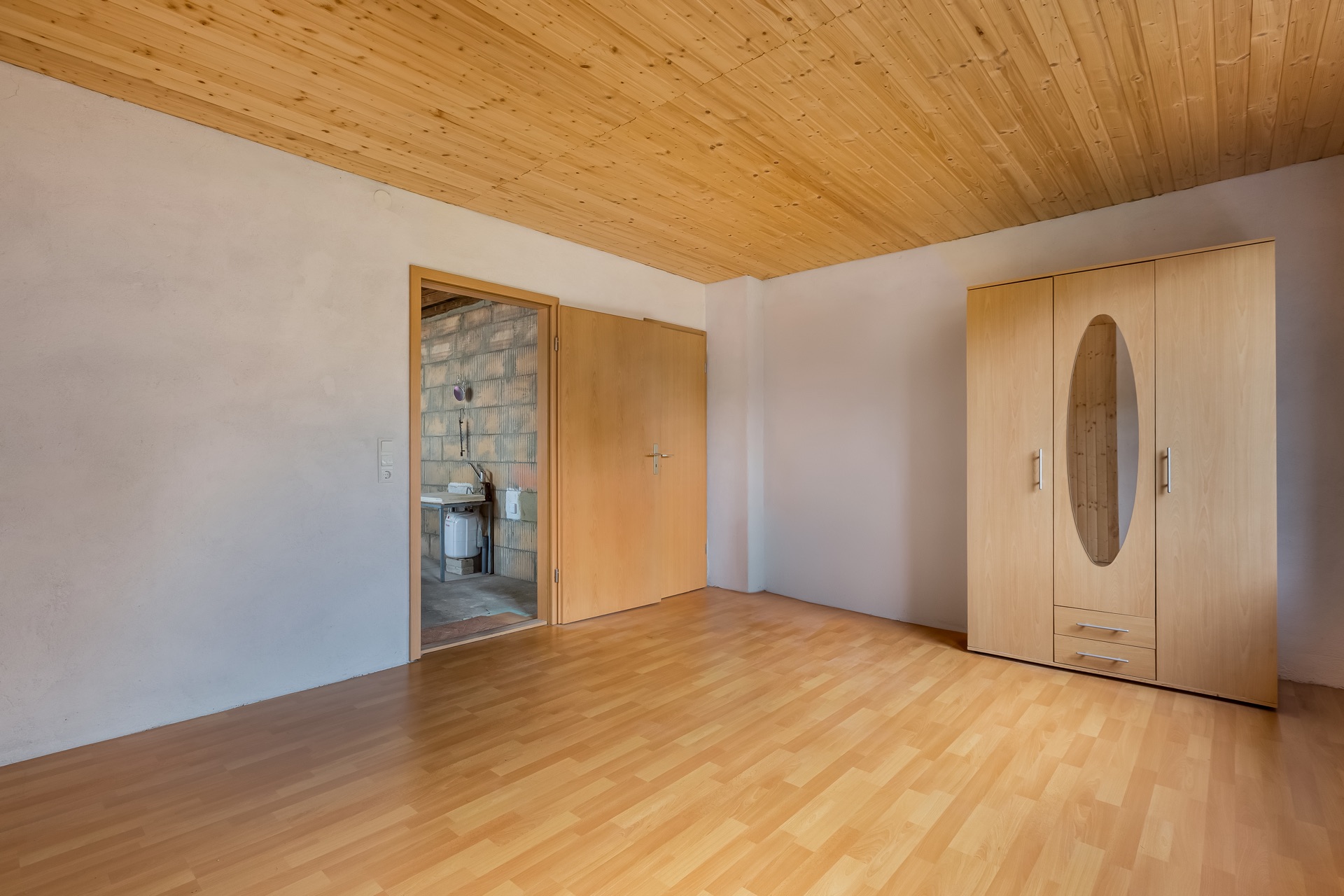 Leeres Zimmer mit Holzdecke, Holzschrank und Blick auf Bad durch offene Tür.