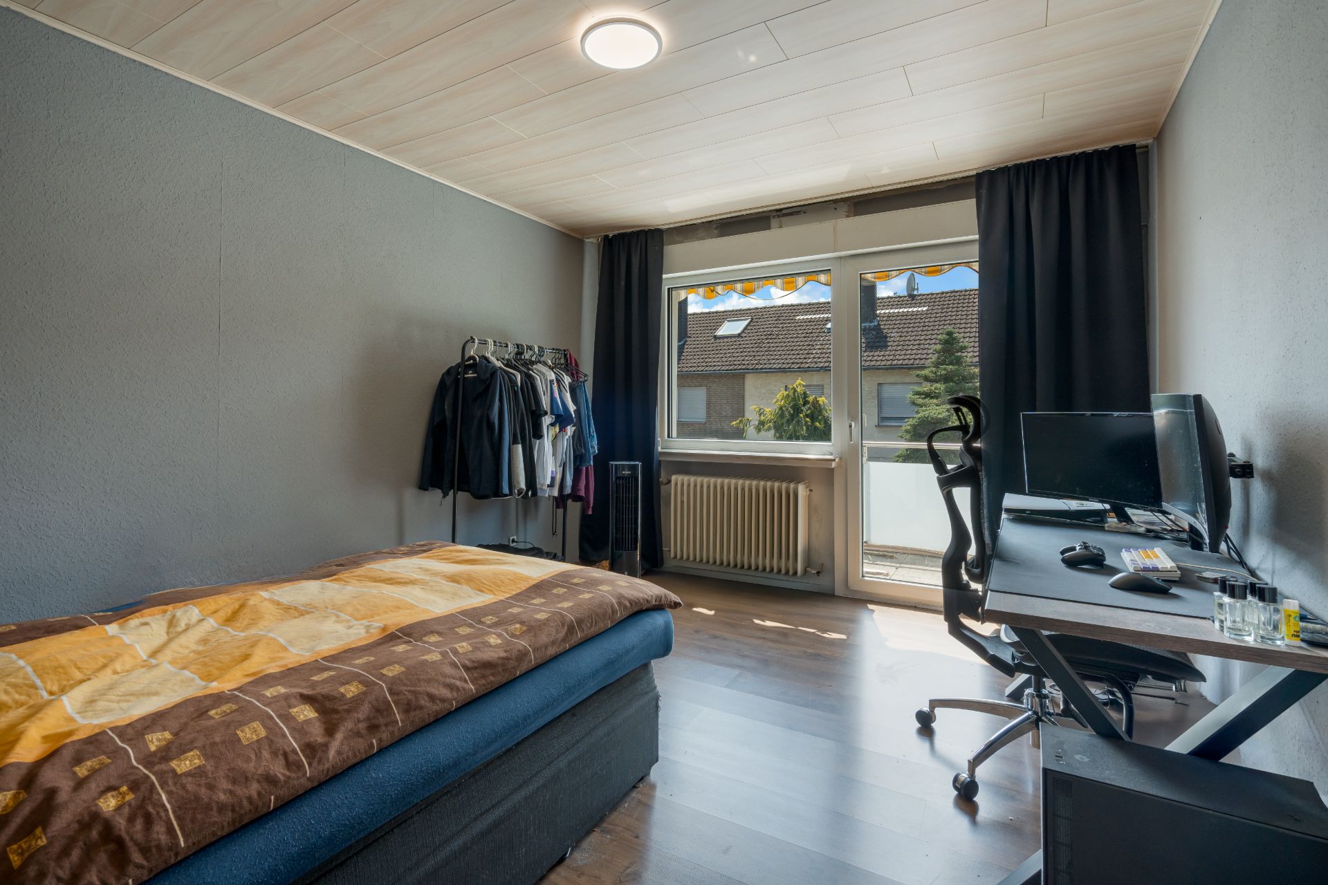Schlafzimmer mit Bett, Schreibtisch, Computer und Kleiderständer vor einem Fenster mit Vorhängen.
