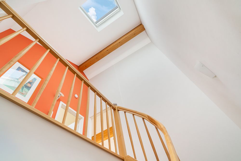 Helles Treppenhaus mit Holzgeländer, Oberlicht und orangefarbener Wand mit Bild.