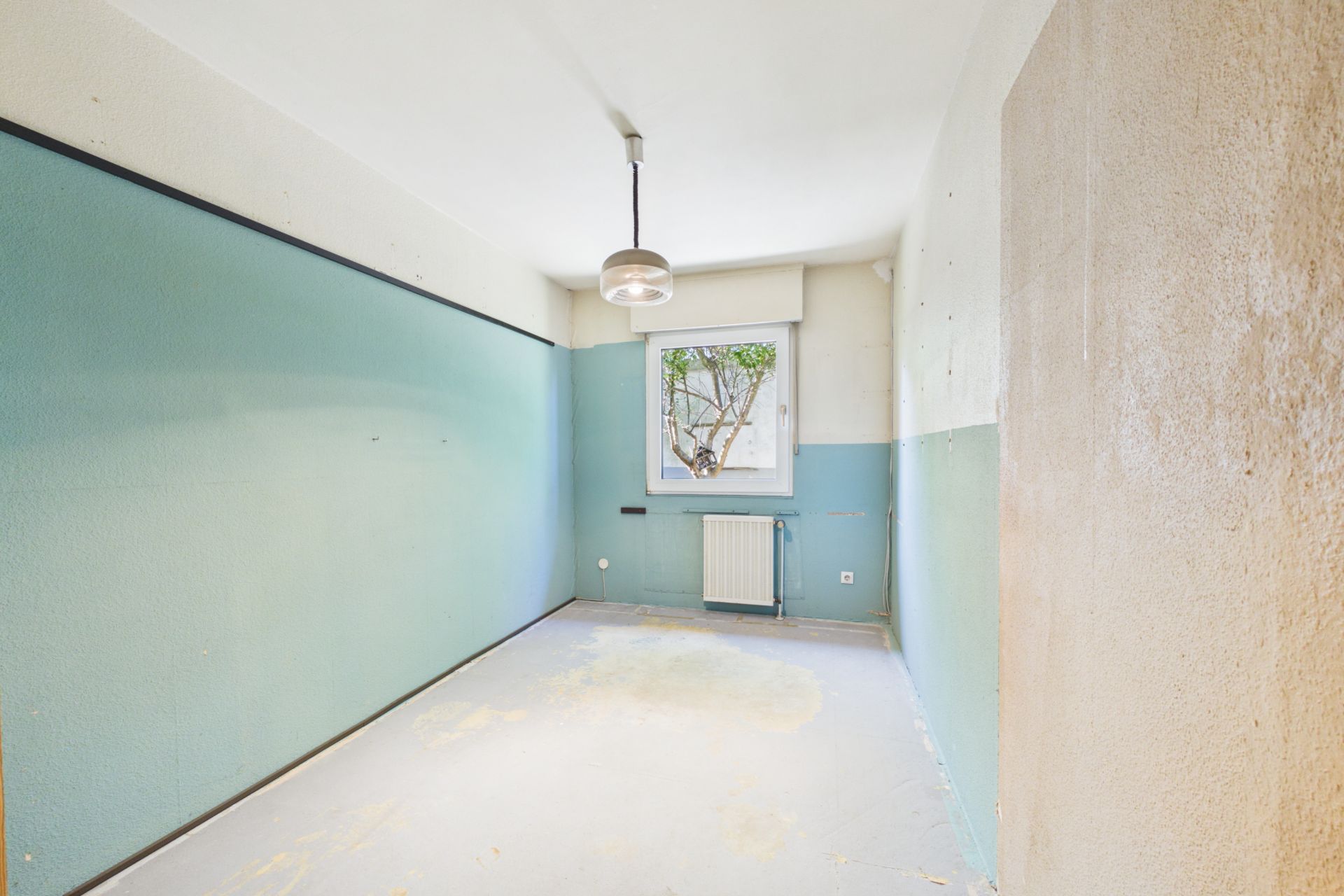 Leerer Raum mit hellblauen Wänden, Fenster, Heizkörper und Deckenlampe.
