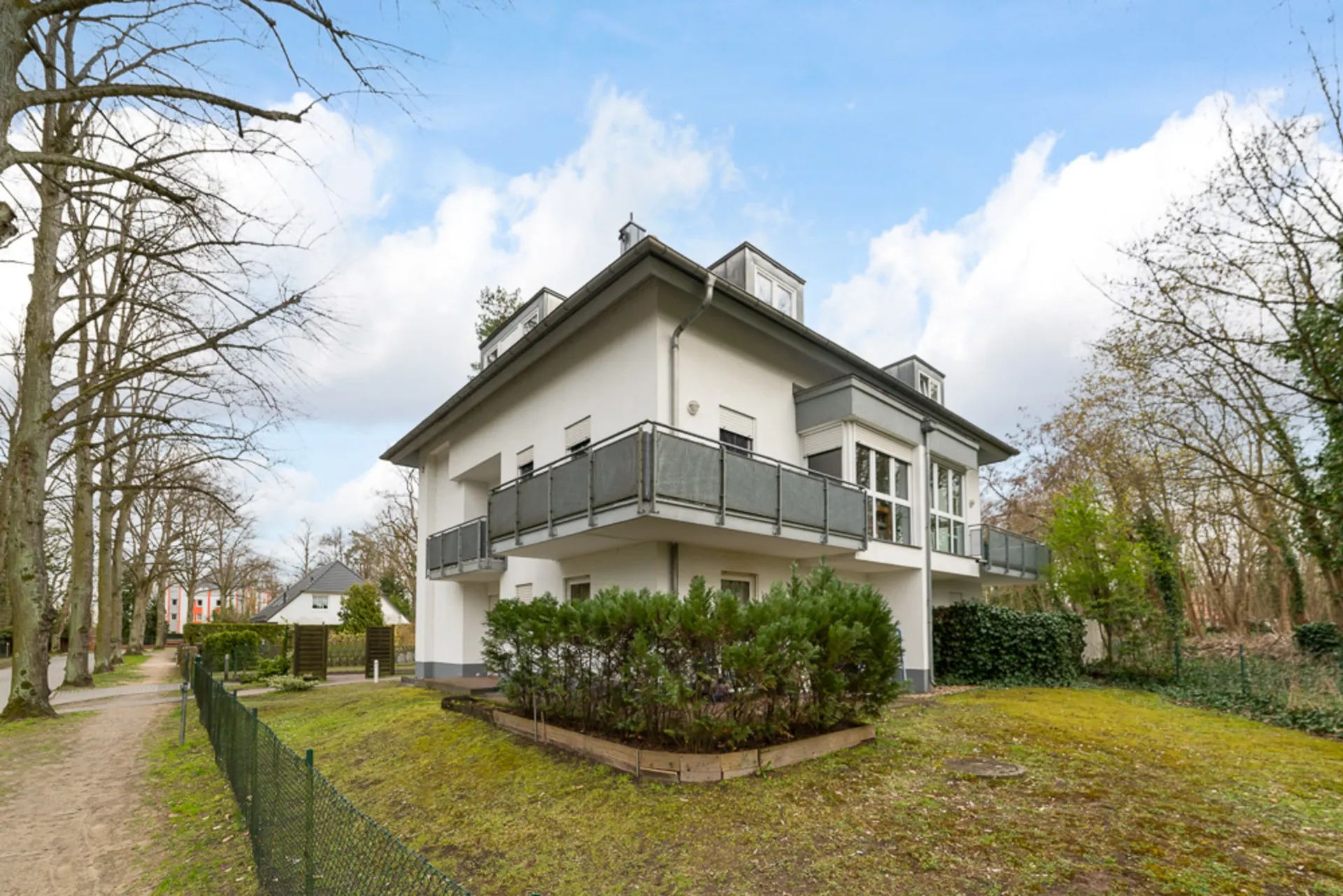 Modernes, weißes Wohnhaus mit Balkon, umgeben von Bäumen und Gartenzaun.