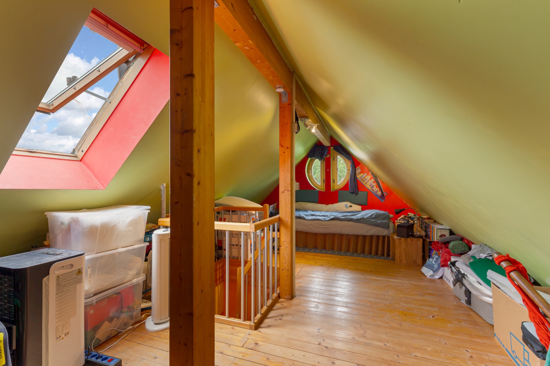 Dachzimmer mit Bett, Holzfußboden, Lagerkisten und Dachfenster mit Blick auf den Himmel.