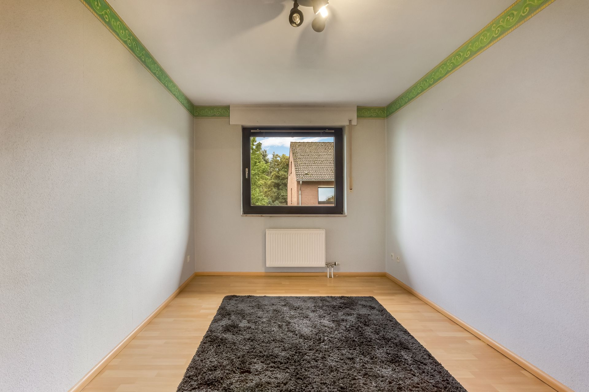 Leeres Zimmer mit Holzfußboden, grauem Teppich und Fenster, durch das ein Haus sichtbar ist.