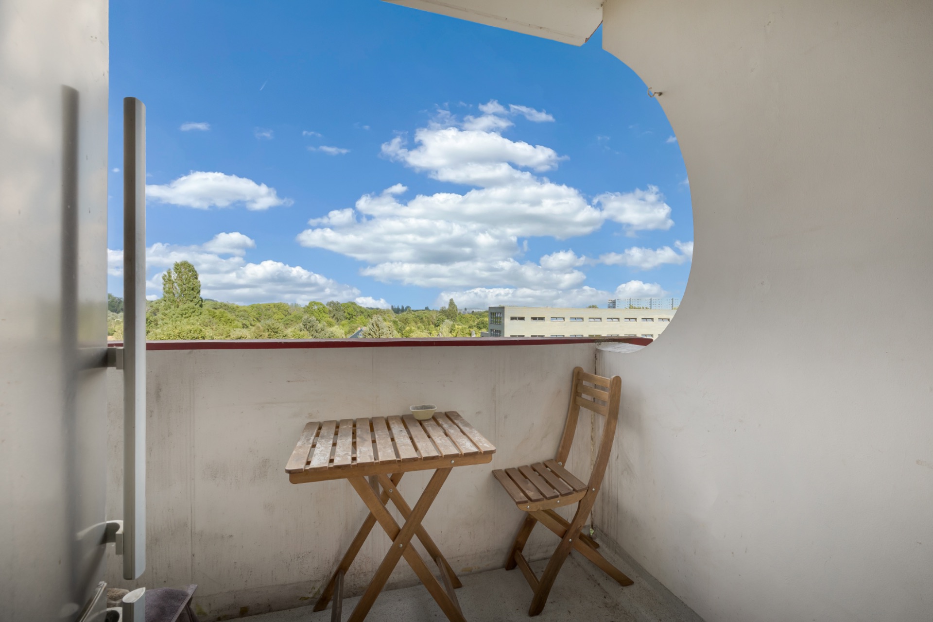 Kleiner Balkon mit Holztisch und zwei Stühlen, Blick ins Grüne unter blauem Himmel.