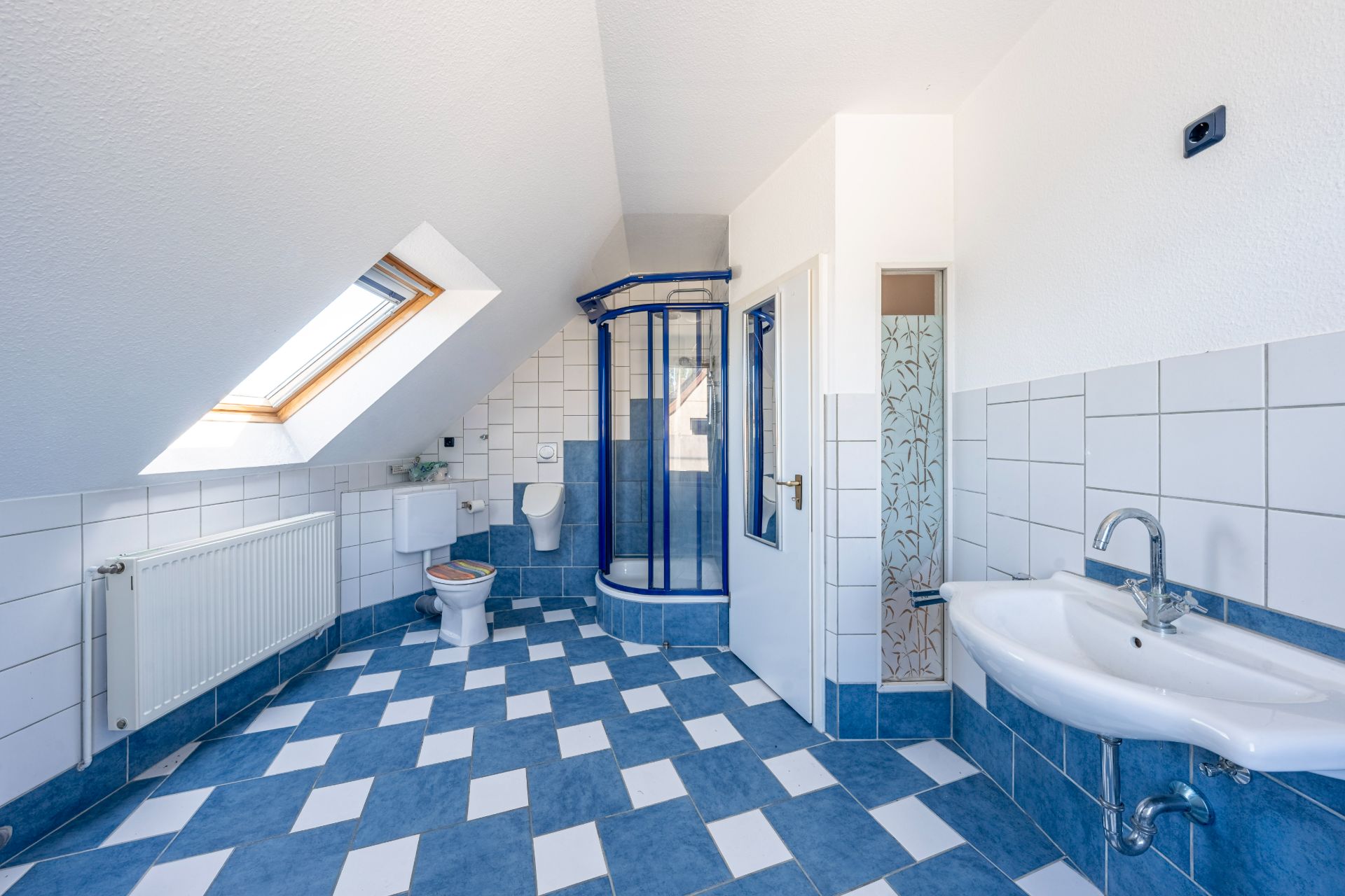 Helles Badezimmer mit blau-weißen Fliesen, Dusche, Waschbecken, Toilette und Dachfenster.