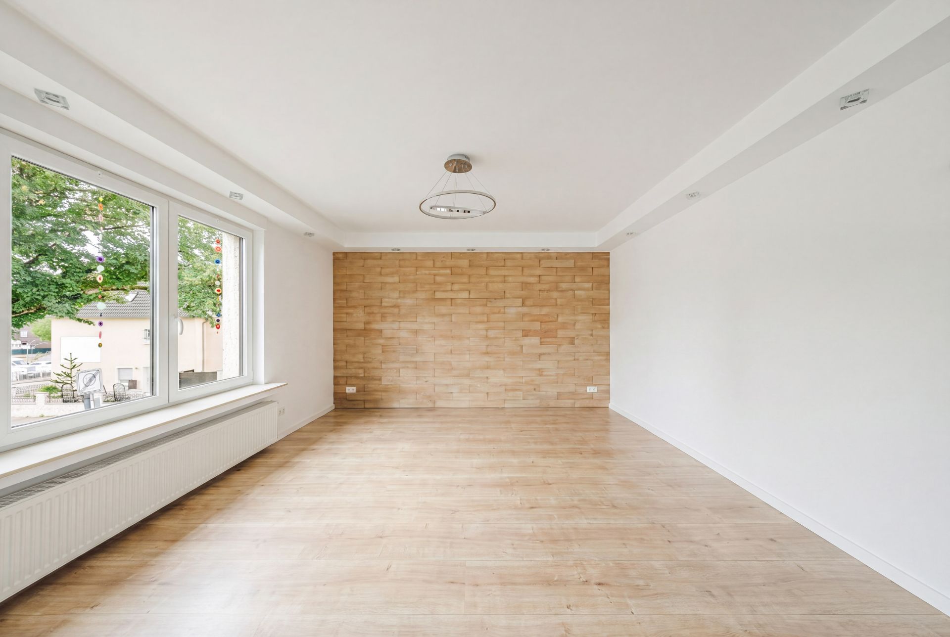 Leerer Raum mit Holzfußboden, heller Wand und großen Fenstern, Wand mit Ziegeloptik.