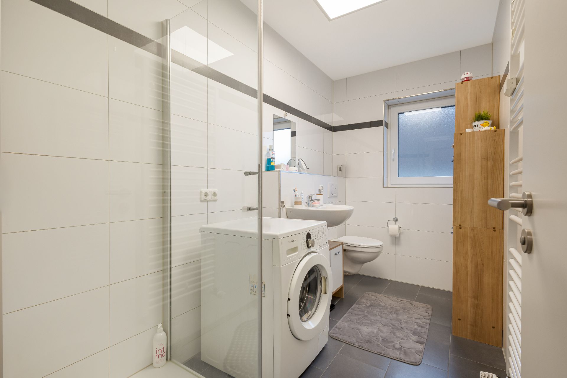 Modernes Badezimmer mit Waschmaschine, Dusche, WC, Waschbecken und Holzregal.