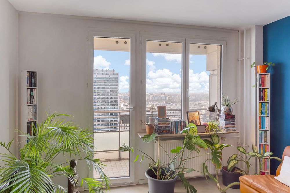 Helles Wohnzimmer mit großen Fenstern, vielen Zimmerpflanzen und Bücherregalen, Blick auf die Stadt.