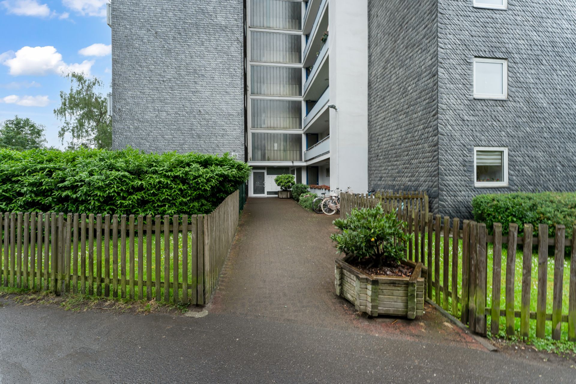 Eingang zu einem Wohngebäude mit Gehweg, Holzzaun und Fahrrädern am Eingang.
