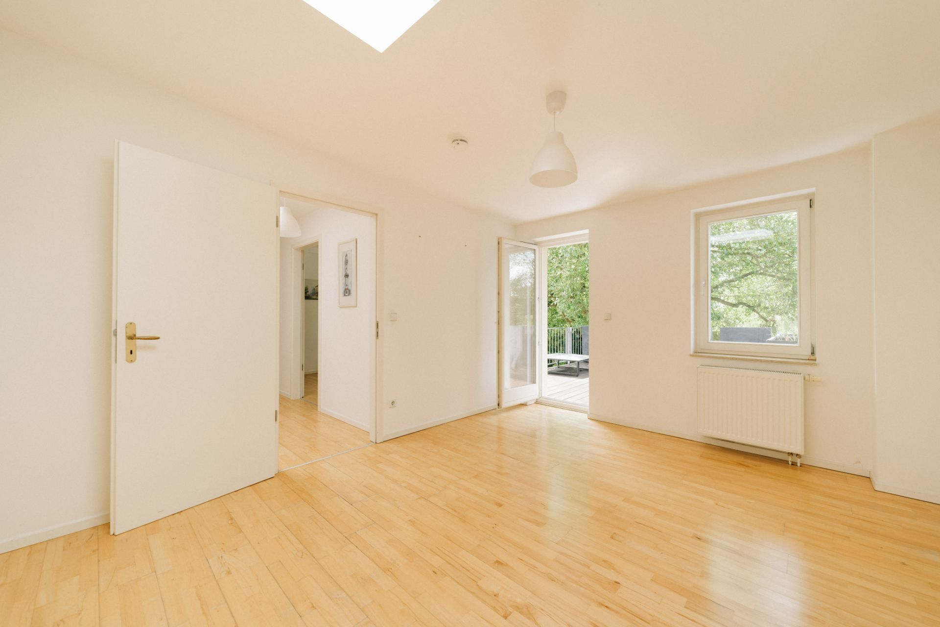 Heller, leerer Raum mit Holzboden, offenem Fenster und Tür zur Terrasse, weiß gestrichene Wände.
