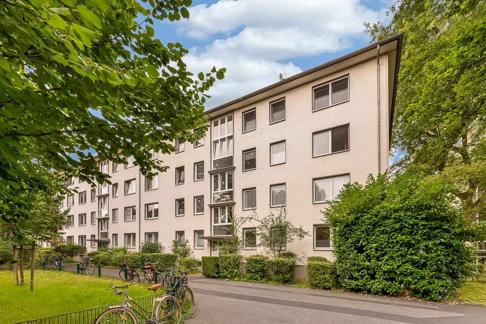 Mehrstöckiges Wohnhaus mit hellen Fassaden, Bäumen, Fahrrädern und Gehweg im Vordergrund.