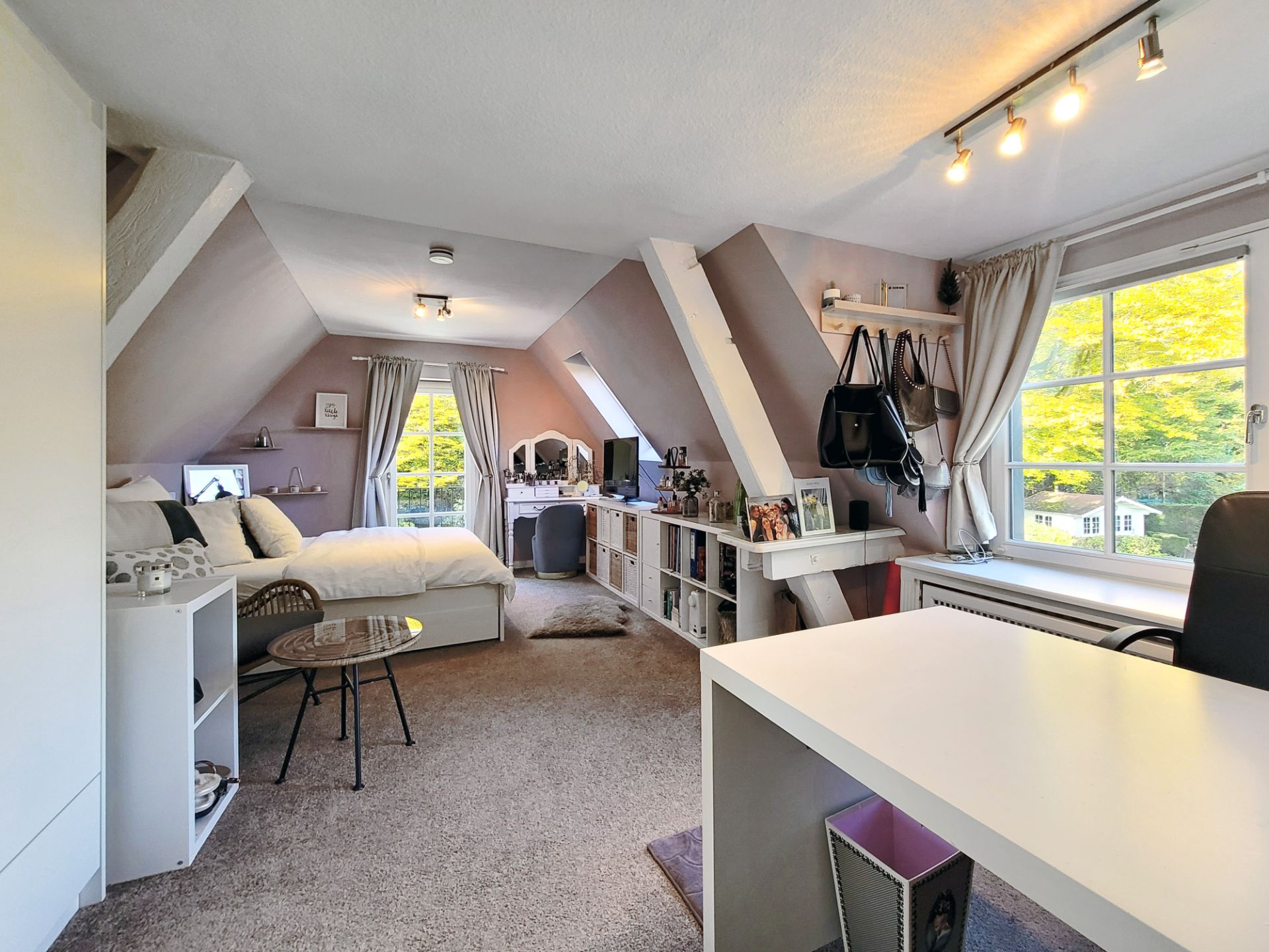 Helles Dachzimmer mit Bett, Schreibtisch, Regalen und großen Fenstern mit Blick ins Grüne.