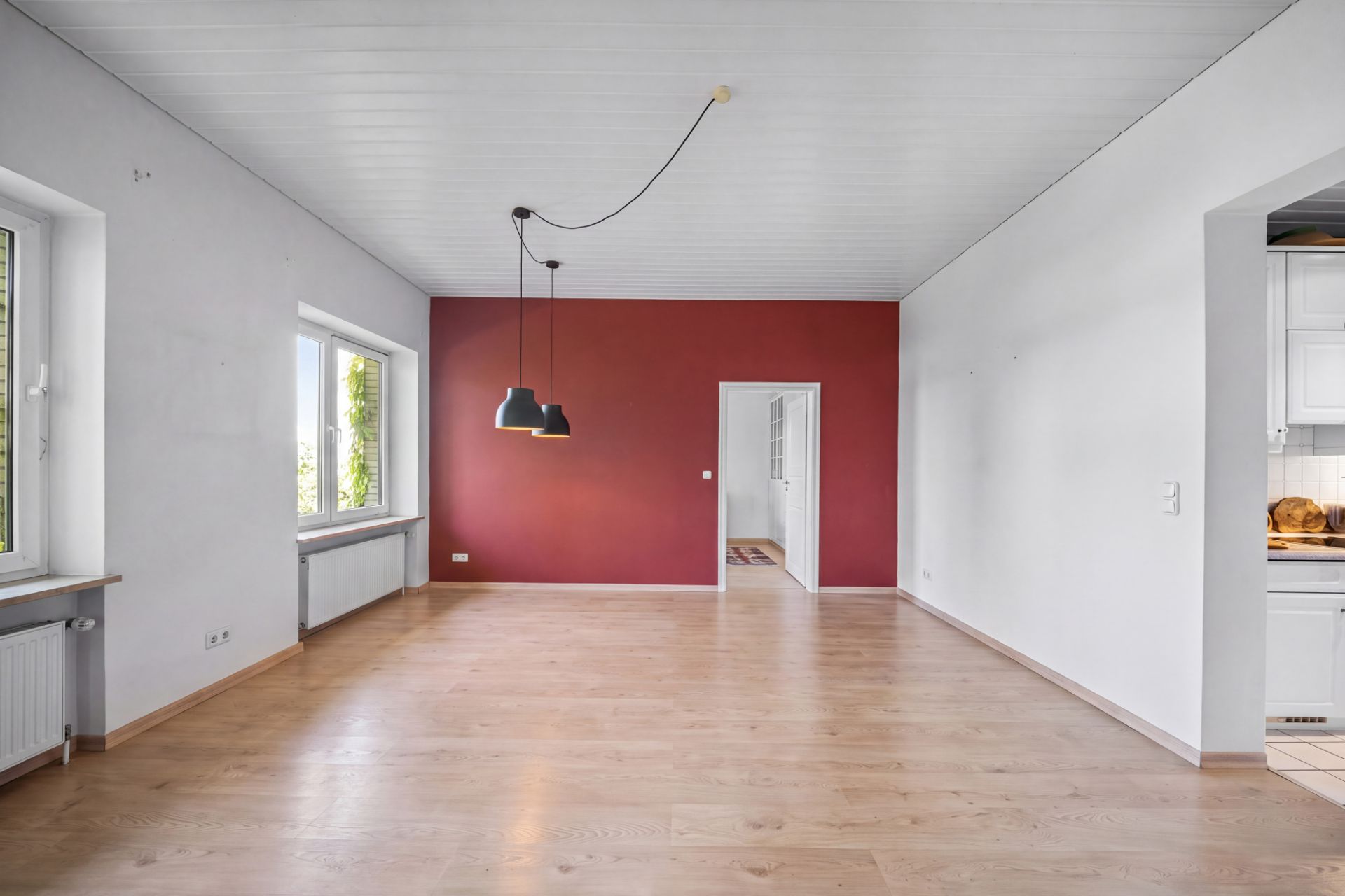 Leeres Wohnzimmer mit Holzfußboden, roten Wand, zwei Fenstern und zwei schwarzen Hängelampen.