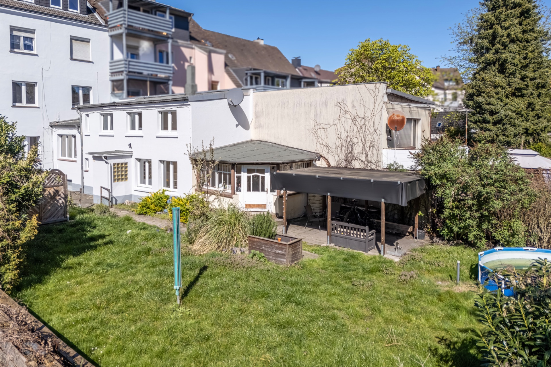 Hinterhof mit Rasenfläche, Terrasse und teilüberdachtem Bereich an einem weißen Wohnhaus.